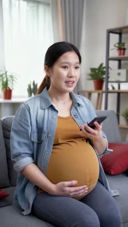 an asian pregnant woman is sitting on a couch holding a mobile phone.