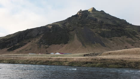 rugged mountain rises above a rural icelandic landscape, with a river flowing in the foreground