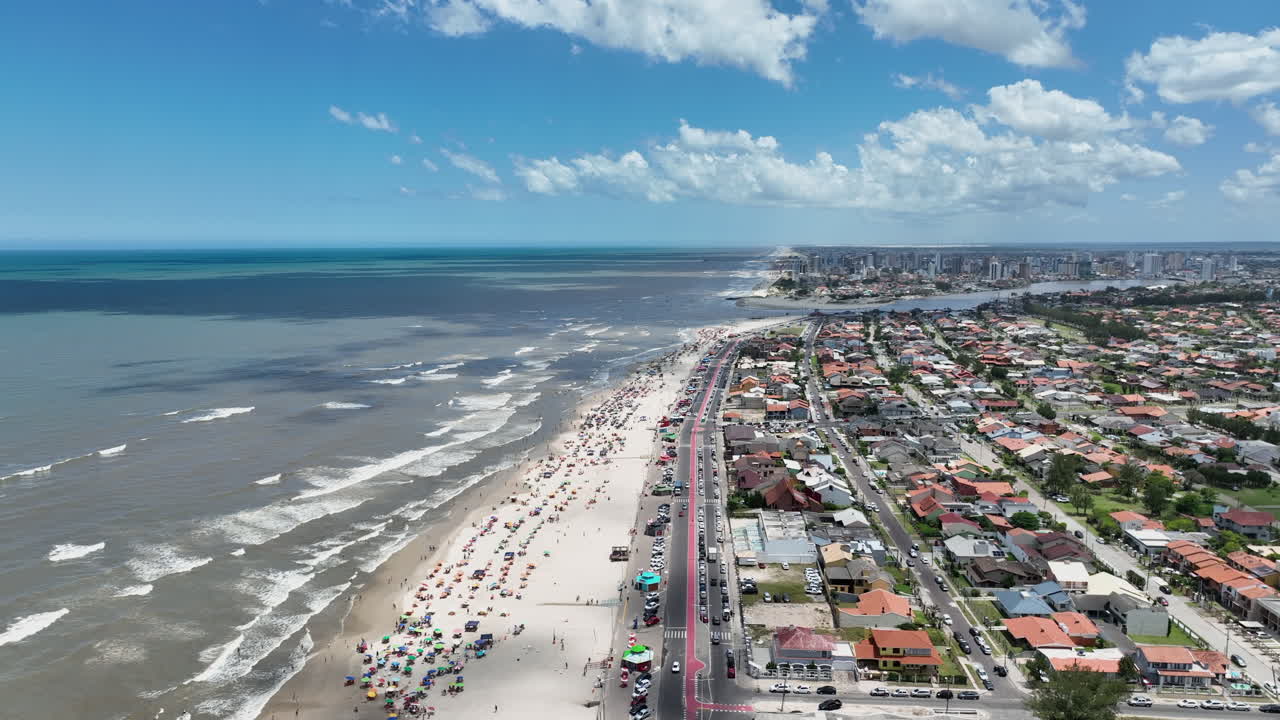 Toma lateral cenital con dron a gran altura con movimiento de cámara siguiendo a un camión, desde la zona urbana continental hacia mar abierto, revelando la extensa y concurrida playa y costa de Imbé, Brasil