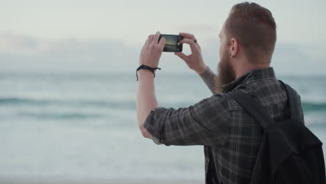 Retrato-De-Un-Joven-Hipster-Usando-Un-Teléfono-Inteligente-Tomando-Una-Foto-De-Un-Tranquilo-Hombre-Caucásico-Barbudo-Junto-Al-Mar-Disfrutando-De-Una-Tranquila-Fotografía-Compartida-En-La-Playa