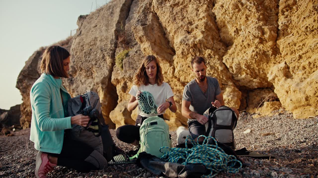 A blonde girl with curly hair in a white T-shirt, a blonde girl with a bob hairstyle in a Blue jacket and a brunette guy in a gray T-shirt are sorting out their backpacks and preparing equipment for rock climbing on a rocky seashore near the yellow rocks on a summer day
