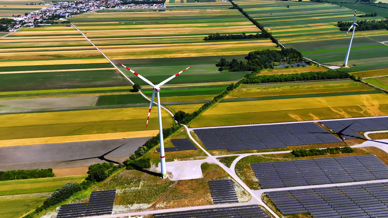 Blades of wind turbine rotate in the wind. Multiple solar panels located around the wind mill. Aerial view