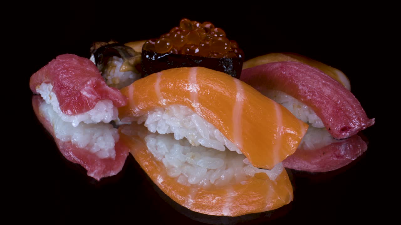 Vista macro di una selezione di nigiri sushi in rotazione su sfondo di vetro nero riflettente: tonno toro, salmone, pesce limone, uova di salmone e anguilla alla brace. Cucina giapponese.