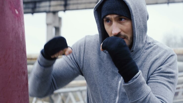 Close-up view of caucasian man in grey hoodie hitting a punching bag outdoors an abandoned factory on a cloudy morning