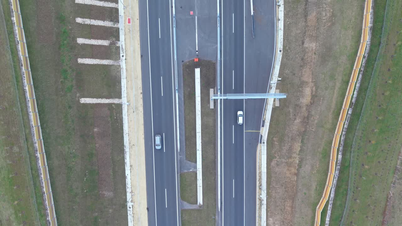 An aerial view of a multilane highway shows cars traveling on separate lanes. The road infrastructure, including barriers, markings, and surrounding grass areas, is clearly visible from above