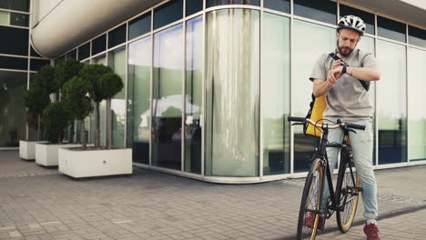 food delivery man wearing thermal backpack stops his bike to take a look at his smartwatch and make sure it is headed to the exact delivery address