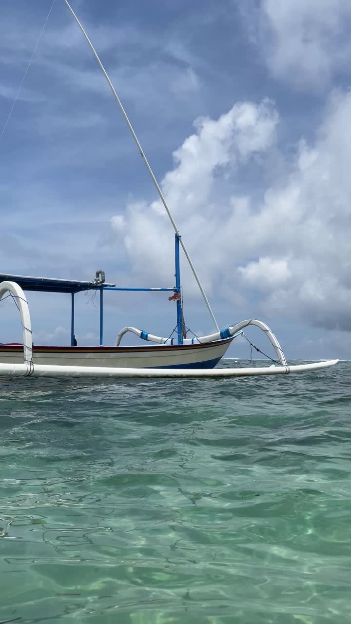 Sociale medier-video, der viser traditionel hvid og blå balinesisk jukung-fiskebåd, der flyder på klart turkisblåt havvand i Nusa Dua, Bali, Indonesien. Roligt hav, åben himmel og kyststemning