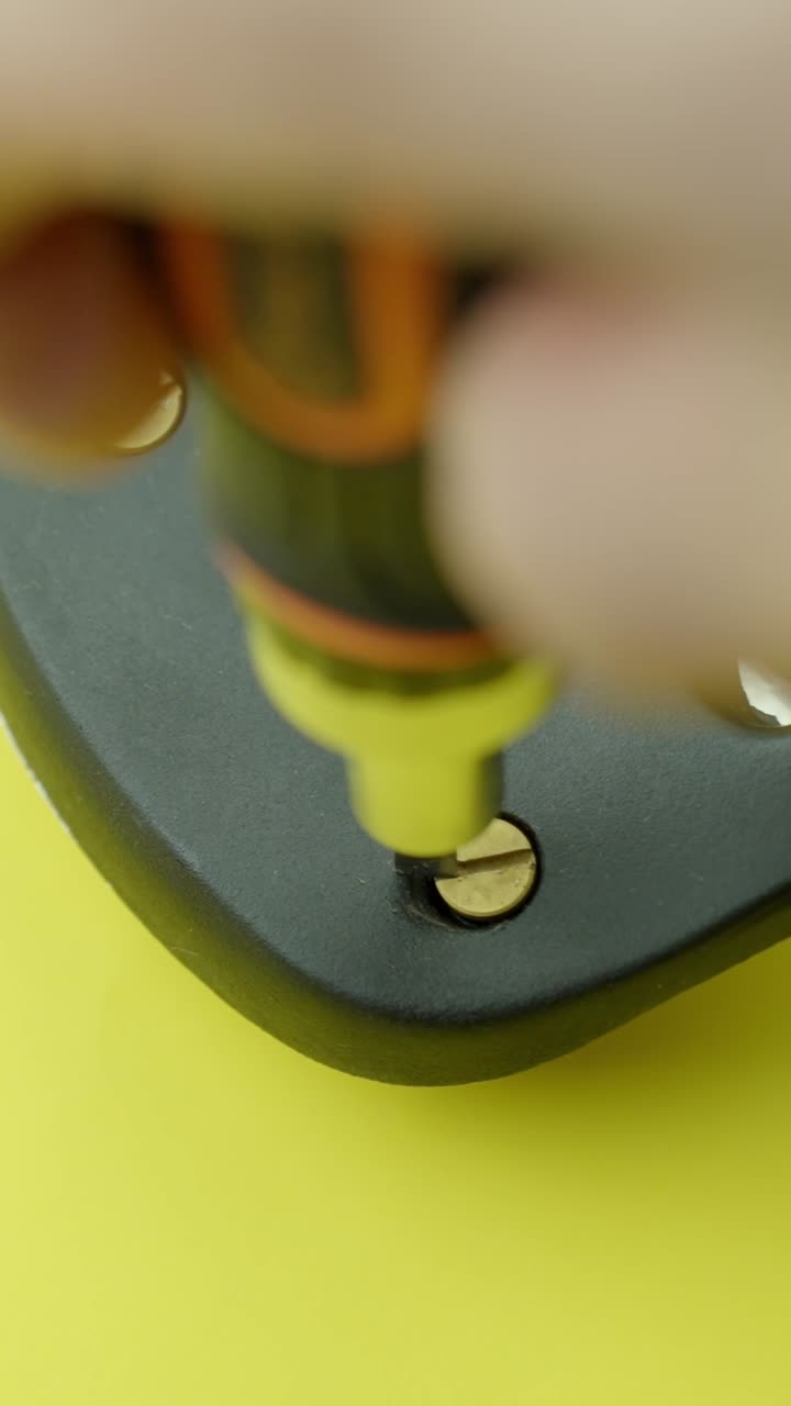 Vertical Macro view of a screwdriver bit tightening a brass screw into a black metal plate against a yellow background. Shot in sharp focus with shallow depth of field