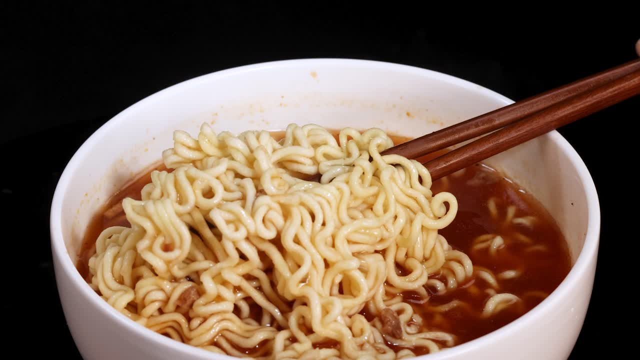 Chopsticks grasp and lift cooked instant noodles from a steaming bowl of broth, captured in close-up with dramatic lighting and a dark background