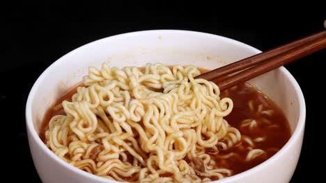 chopsticks grasp and lift cooked instant noodles from a steaming bowl of broth, captured in close-up with dramatic lighting and a dark background
