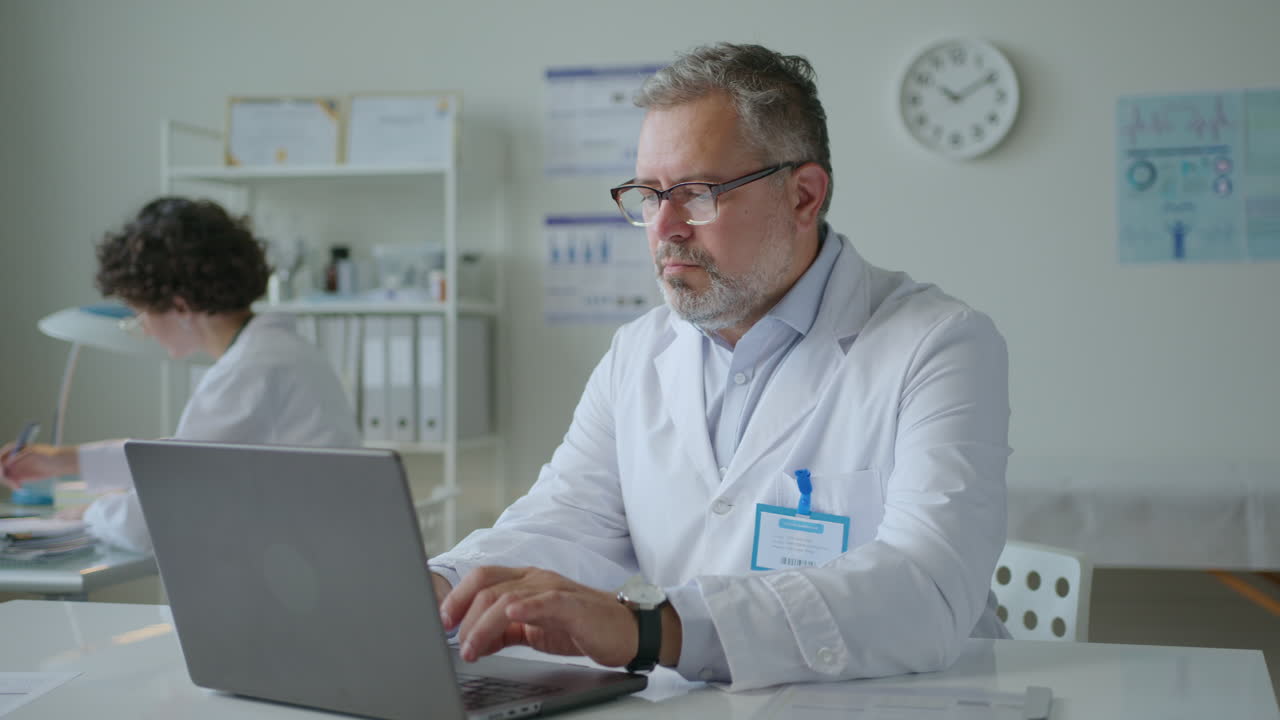 Senior Doctor Working on Laptop at Desk in Clinic