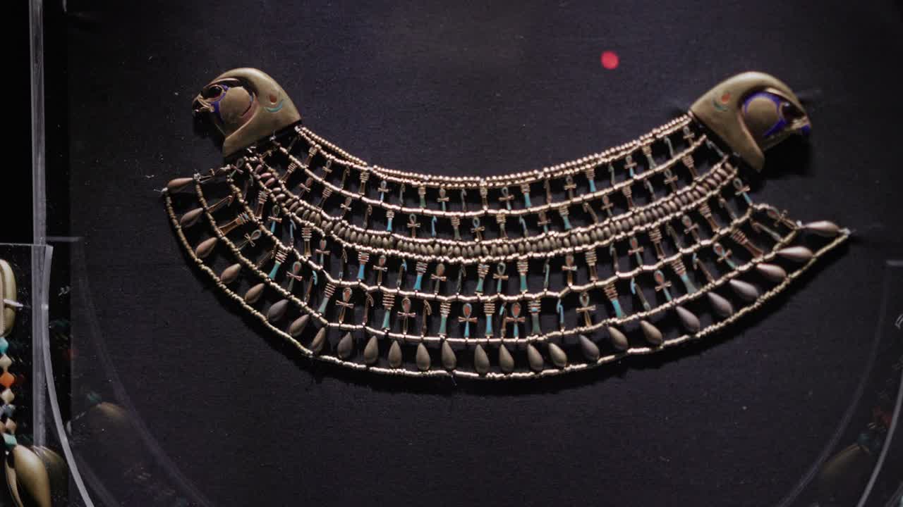 Primo piano di una collana a collare larga dell'antico Egitto esposta in una teca museale, caratterizzata da intricate perline, file di motivi simbolici ankh e terminali stilizzati a testa di falco.