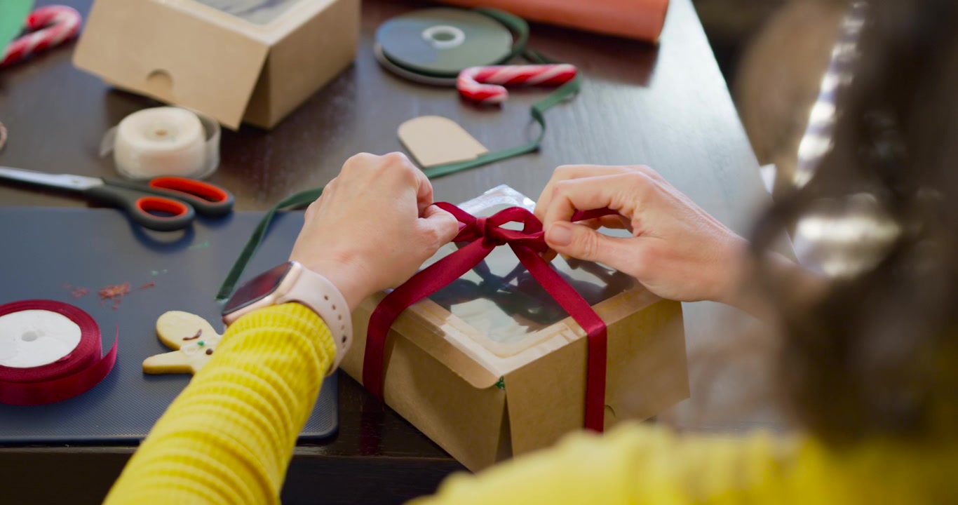 Woman pulling and arranging red ribbon, tying bow to finish windowed gift box in yellow top