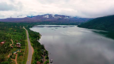 Vista-Aérea-Por-Drones-De-Un-Hermoso-Lago-De-Montaña-Con-Latas-Rodantes-En-El-Fondo-Mientras-Los-Autos-Conducen-Por-La-Carretera-De-Colorado-A-Lo-Largo-De-La-Costa-De-La-Montaña-De-Sombra-Y-El-Gran-Lago