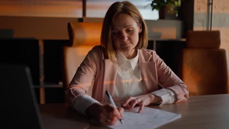 Portrait-of-a-confident-middle-aged-blonde-girl-in-a-pink-jacket-and-modern-uniform-who-sits-at-a-wooden-table-in-a-sunny-office-and-smiles