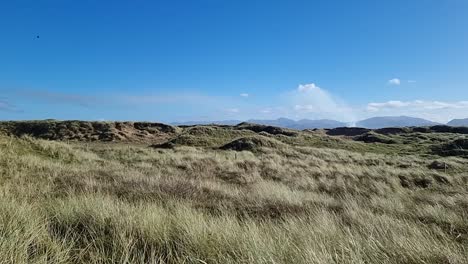 Slow-motion-panning-across-grassy-sand-dune-landscape-with-hazy-Snowdonia-mountain-range-on-sunrise-horizon