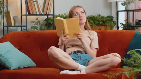 young woman reading interesting book turning pages smiling enjoying literature taking rest on couch