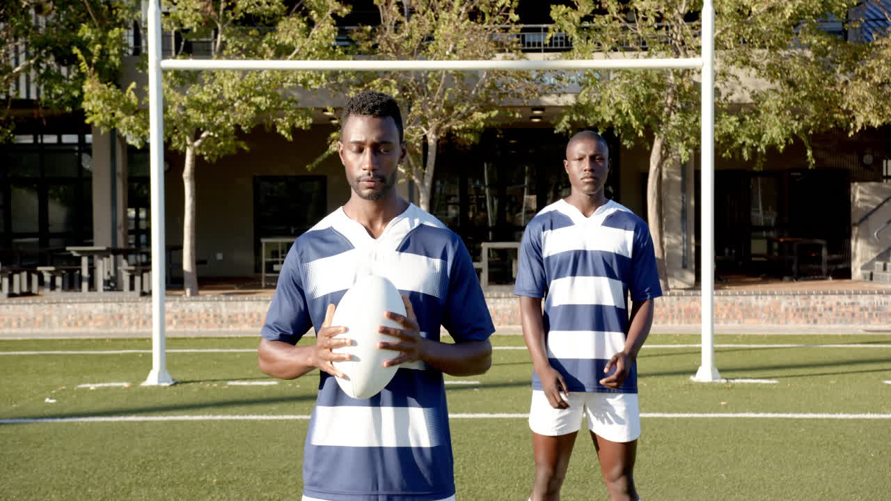 Holding rugby ball, two african american men in sports uniforms standing on field, copy space