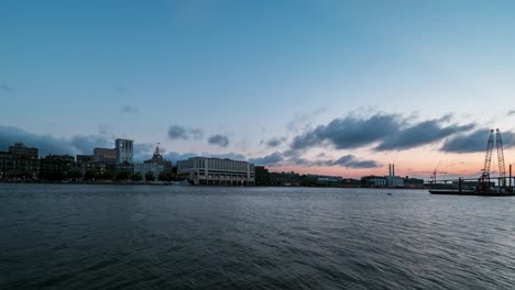 Tag-zu-Nacht-Zeitraffer-Der-Skyline-Von-Savannah-Georgia-über-Dem-Savannah-River,-Während-Boote-Und-Frachtschiffe-Vorbeifahren