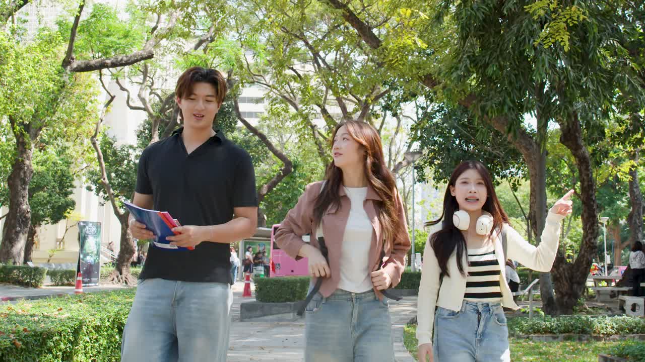 นักศึกษาชาวเอเชียเดินผ่านสวนสาธารณะในวิทยาเขตที่แดดจ้า ถือสมุดโน้ตและสวมชุดยีนส์ลำลอง