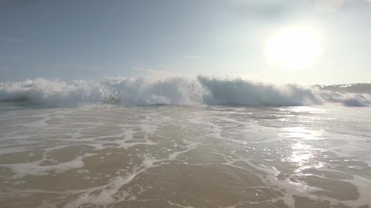 A sunlit Indian Ocean wave advances through shallow surf and breaks directly over the waterline camera, engulfing the lens in whitewater and spray at Mirissa Beach, Sri Lanka.