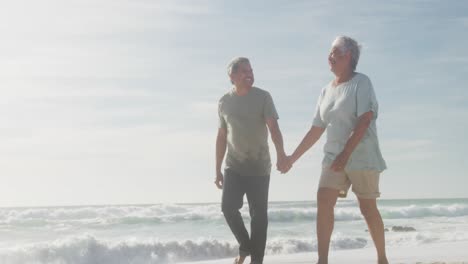 Feliz-Pareja-De-Ancianos-Hispanos-Tomados-De-La-Mano,-Caminando-Por-La-Playa-Al-Atardecer