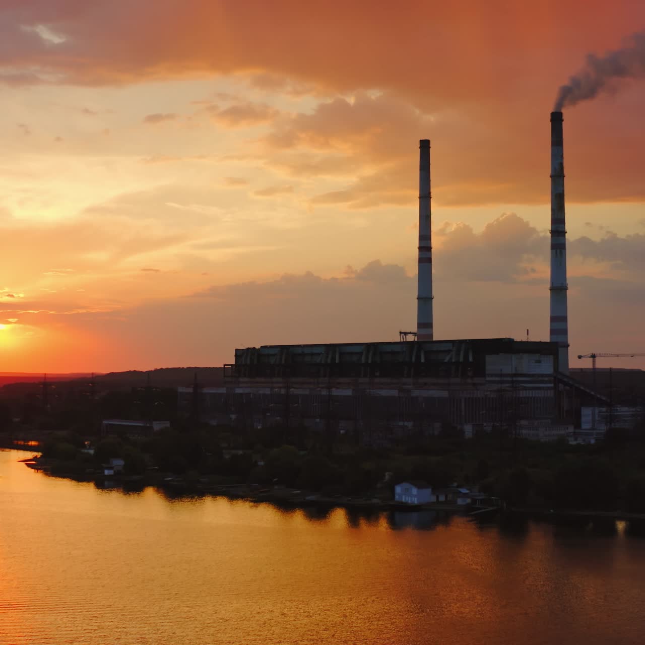 Factory on orange background. Harmful chemical plant over the river at sunset. Large territory of industrial plant in the countryside
