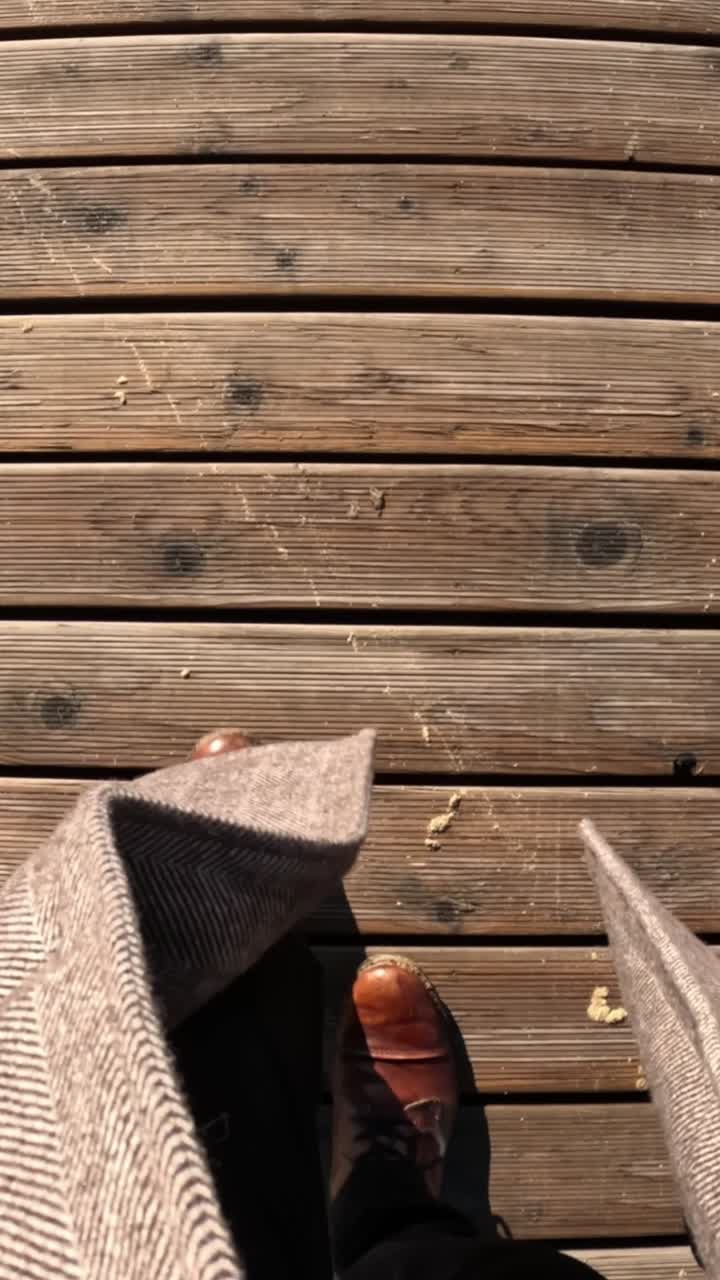 First-person view of walking across a sunlit wooden boardwalk wearing brown leather shoes and black pants.