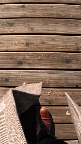 first-person view of walking across a sunlit wooden boardwalk wearing brown leather shoes and black pants.