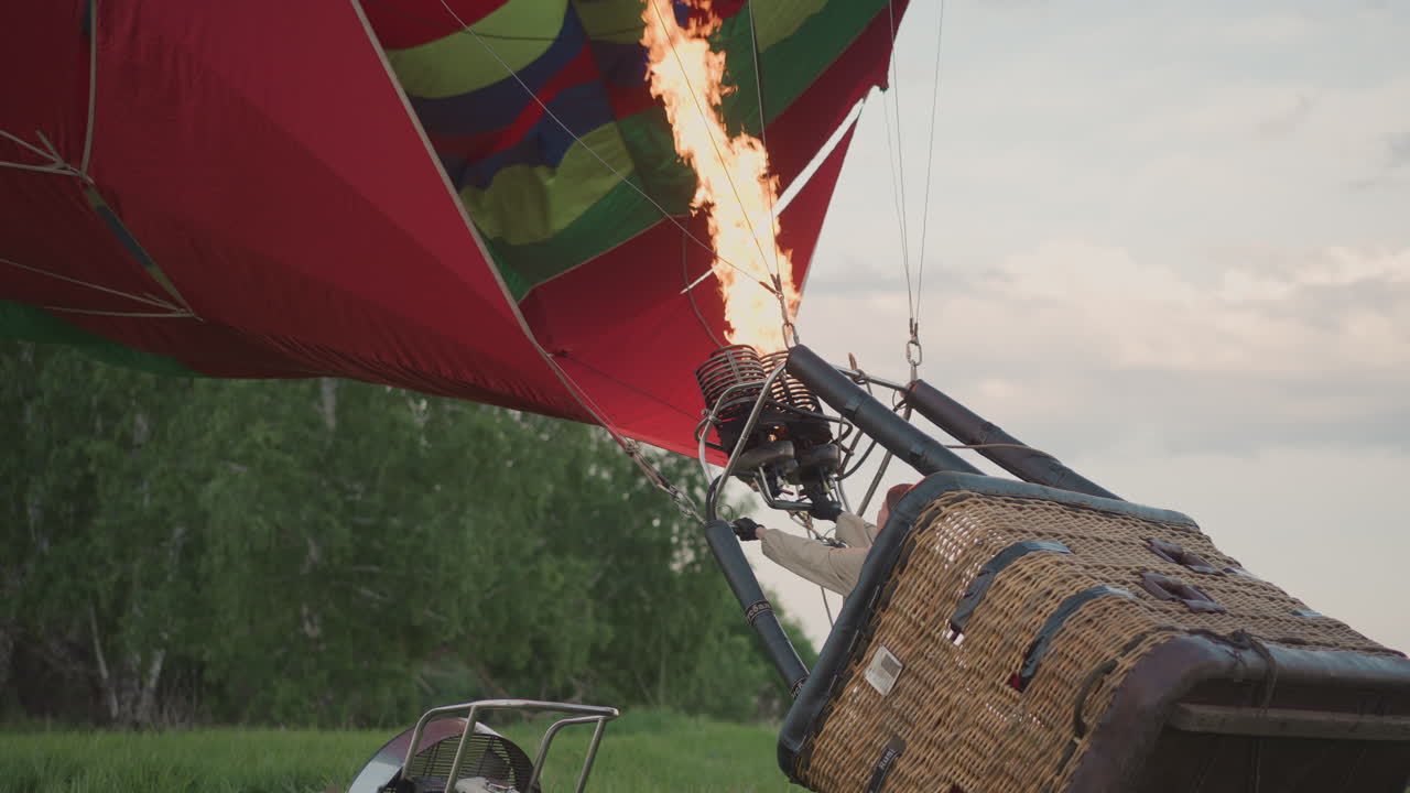 pilot igniting burner to inflate hot air balloon envelope in open field at sunset with basket and fan in view while holding ropes taut under twilight sky during preflight launch procedure