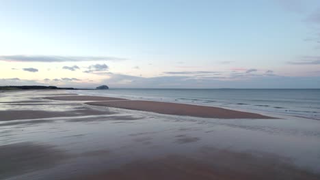 a drone facing out across the calm ocean slowly turns left to reveal a subset reflecting off of a sandy beach