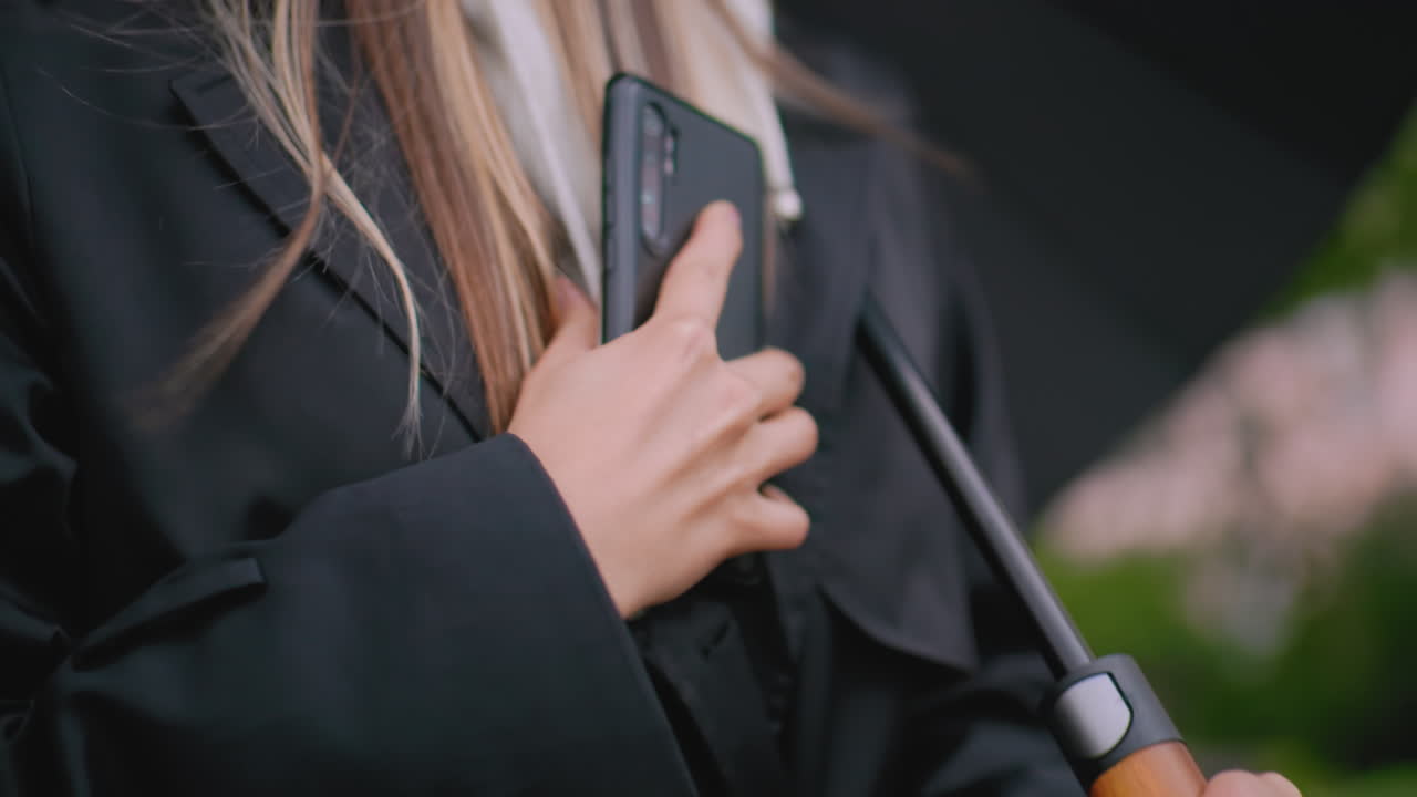 Close up of young woman wearing black coat holding smartphone and umbrella outdoors on rainy day, showing hand grip and modern device with blurred natural green background