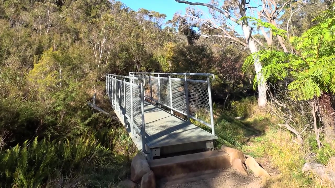 Kamera se pohybuje směrem ke kovové lávce pro pěší na slunné turistické stezce v Austrálii.