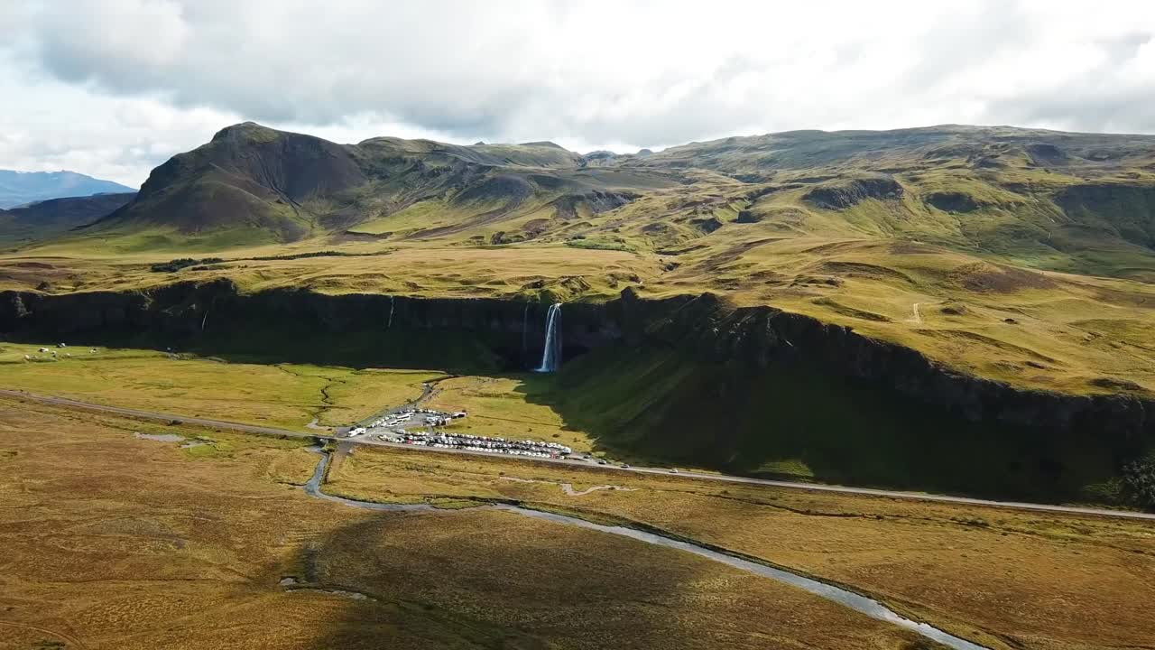Seljalandsfoss waterfall flows dramatically from cliffs in Iceland's rugged terrain, surrounded by golden plains, green hills, and picturesque natural beauty under a partly cloudy sky.