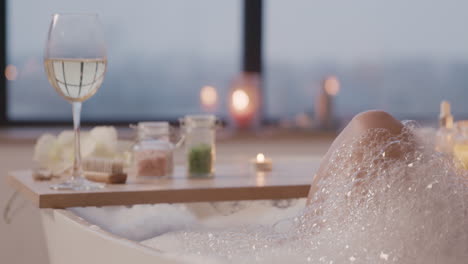 Camera-Focuses-On-A-Wooden-Board-Above-A-Bathtub-With-A-Glass-Of-Wine,-Bath-Salts-And-A-Candle