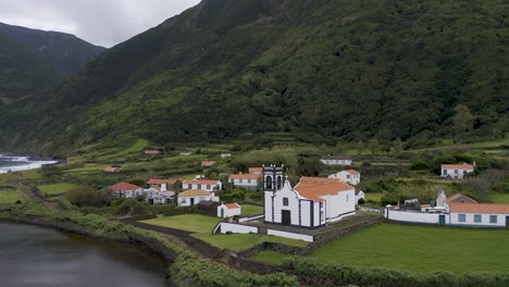 drone orbiting view of a coastal village church, lush green cliffs landscape, fajã de santo cristo, são jorge island, the azores, portugal