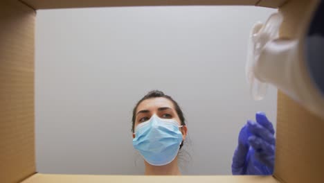 Woman-in-Mask-Taking-Cleaning-Supplies-From-Box.home-delivery,-shipping-and-pandemic-concept-woman-in-protective-medical-mask-and-gloves-opening-parcel-box,-looking-inside-and-taking-cleaning-supplies-out