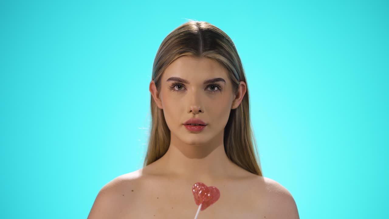 Static shot of a young caucasian naked woman licking a heart shaped lollipop while looking sexy into the camera in slow motion against turquoise background