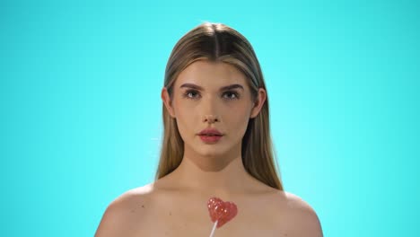 static shot of a young caucasian naked woman licking a heart shaped lollipop while looking sexy into the camera in slow motion against turquoise background