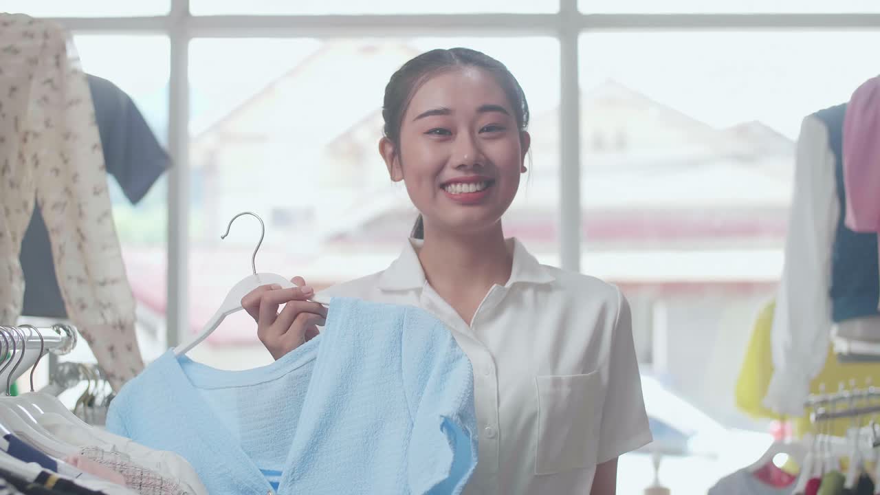 젊은 아시아 여성이 파란색 옷을 선택하고 미소 짓고 부티크에서 쇼핑하는 동안 카메라에 신용카드를 보여줍니다.