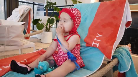 female toddler sitting on a poolside chair and putting blue strap in her mouth