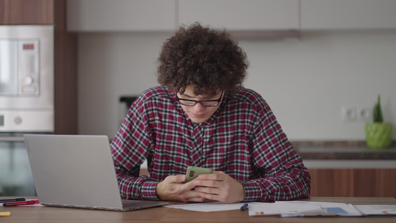 Curly haired Male student attractive young boy in glasses is studying at home using laptop typing writing in notebook. College student using laptop computer watching distance online learning seminar