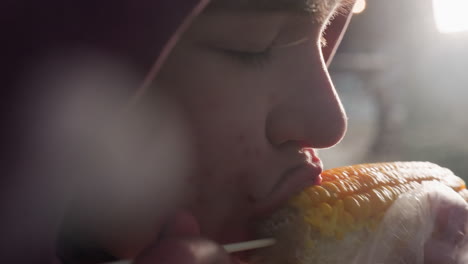 close-up of person eating corn held by stick wrapped in white plastic with bokeh lights in blurred background, steam rising from fresh corn, enjoying tasty outdoor snack