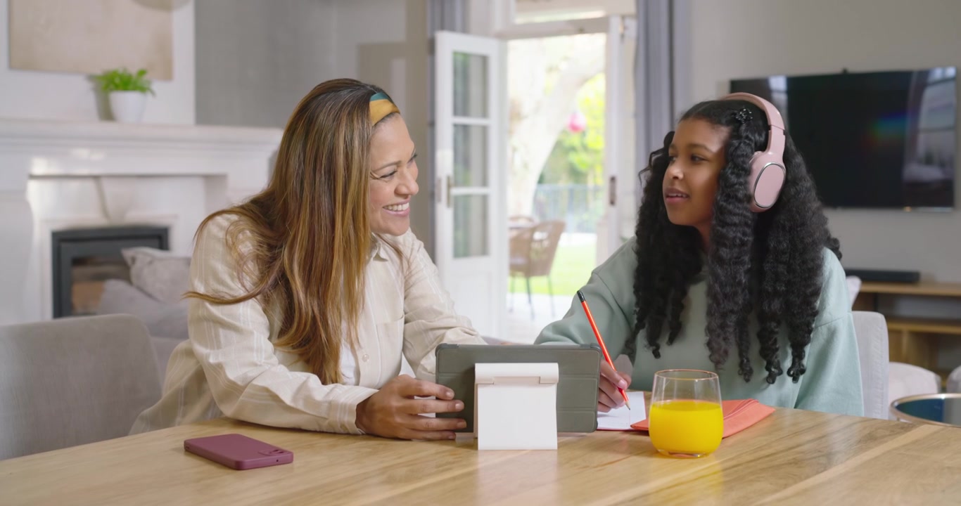 Mamma e figlia adolescente afroamericane che studiano a casa, la mamma che indica una lezione sul tablet mentre l'adolescente scrive.