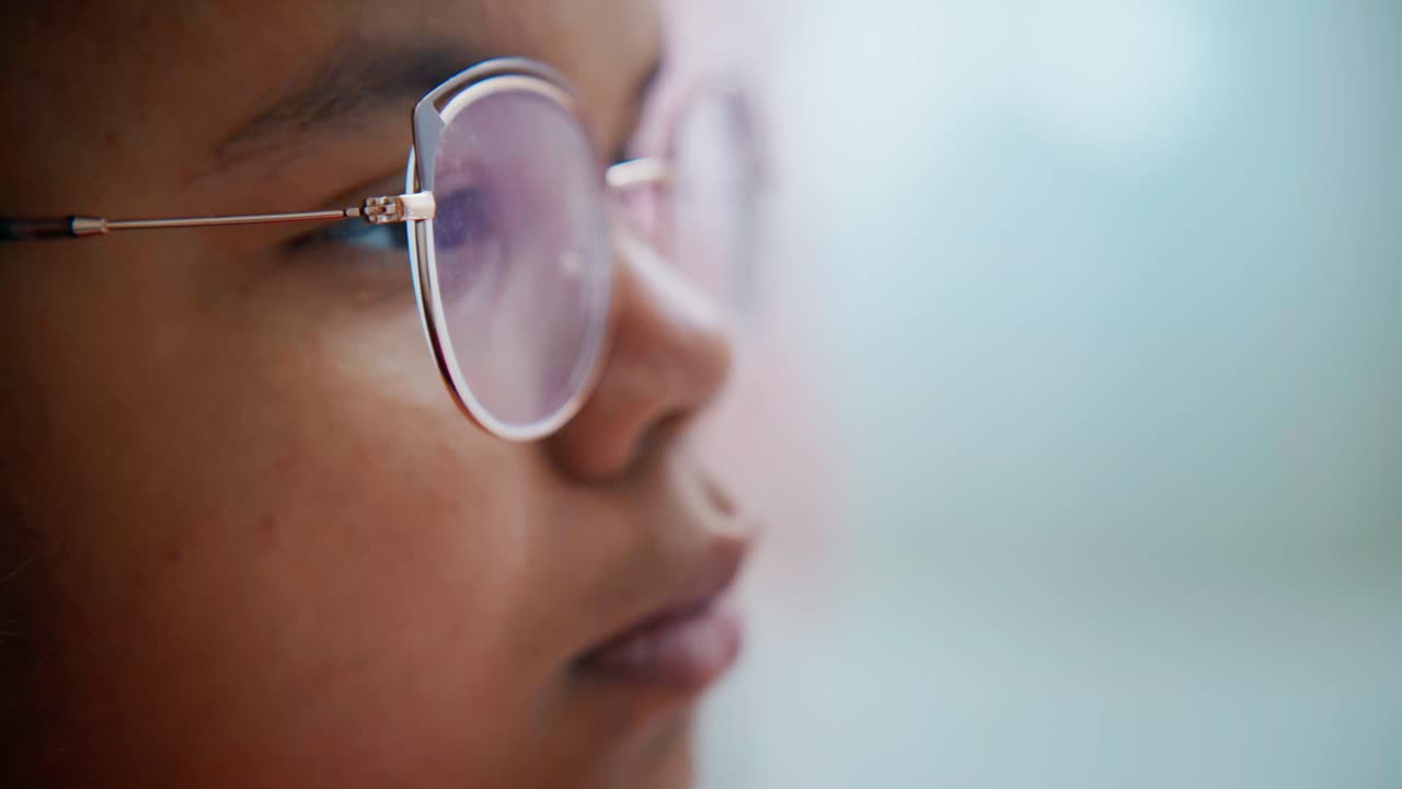 Close up of a young girl wearing round metal rimmed glasses, possibly a student, engrossed in reading, her face showing concentration and focus