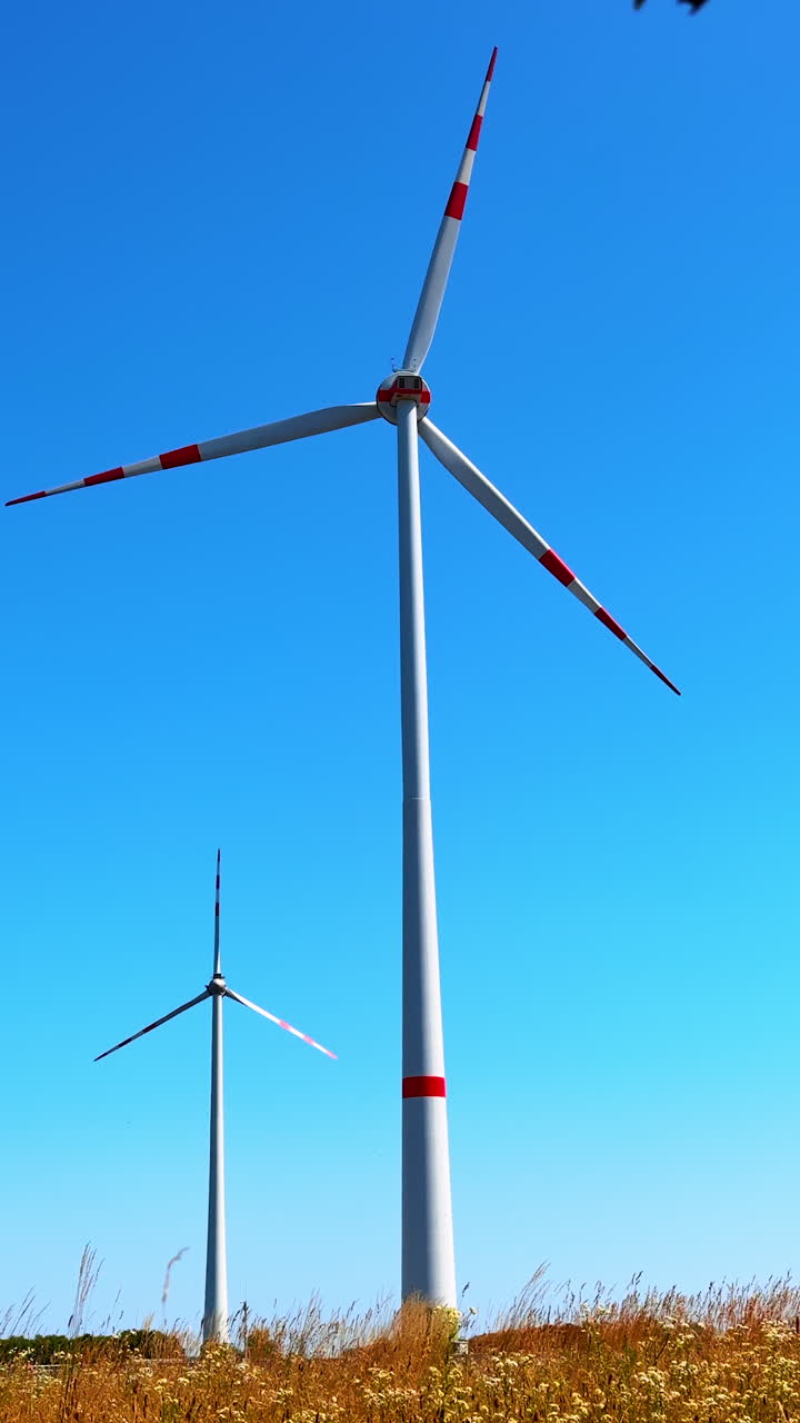 Two wind turbines rotating at the backdrop of blue clear sky. Low angle view at the windmills producing clean energy. Vertical video.