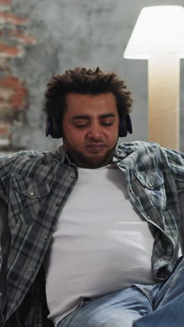 Enraptured black guy with earphones plays imaginative musical instrument resting on foiled sofa. African-American man at break during restoration work