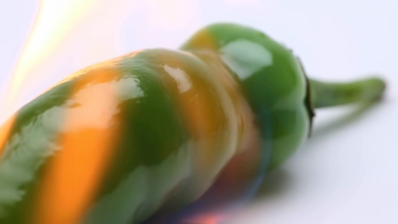 Primo piano di peperoncino verde che brucia a fiamma viva, mostrando un vivido cambiamento dalla pelle liscia e lucida a una carbonizzazione vescicolosa e annerita, texture bollente, fumo, calore e drammatico dettaglio culinario