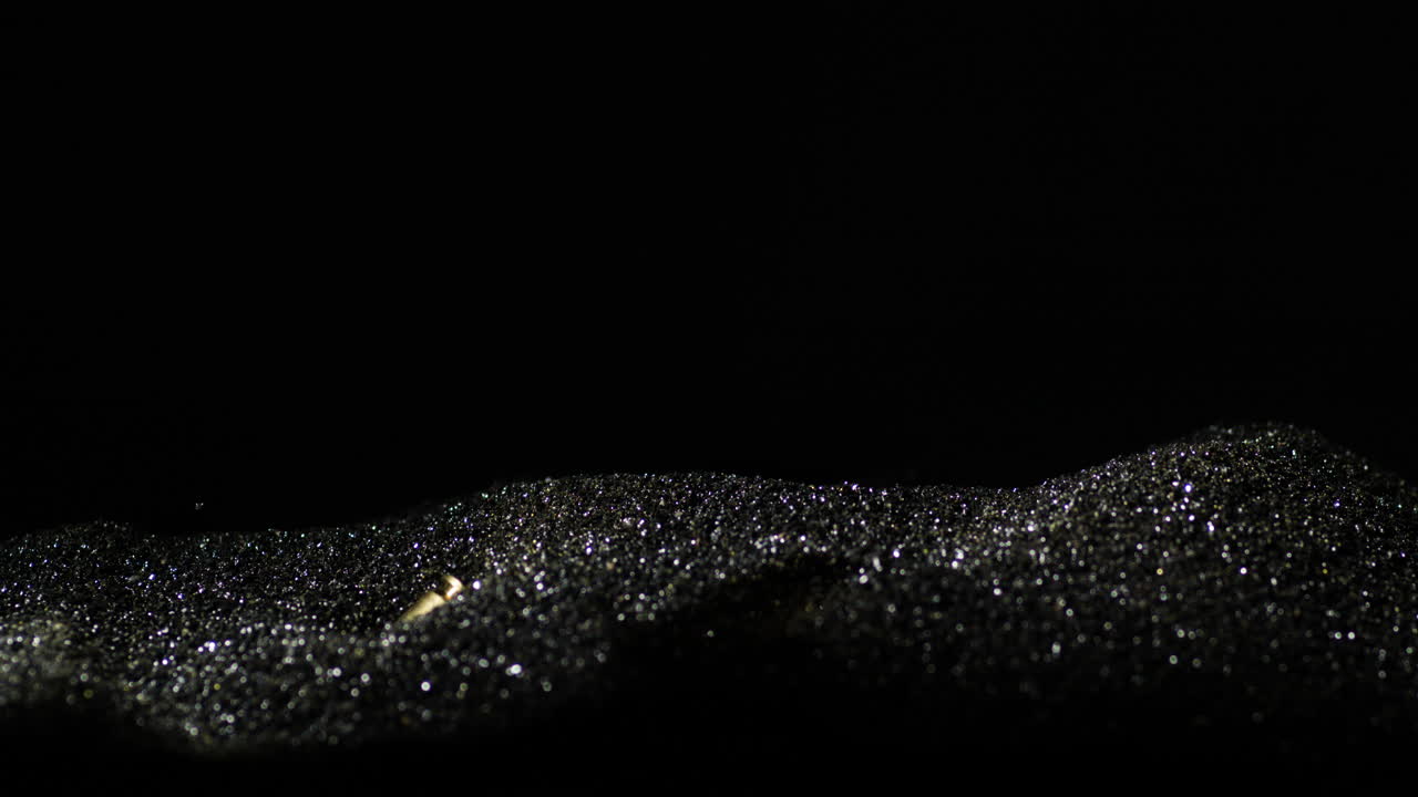 Proiettili d'oro da 9mm che cadono su sabbia nero-argento con sfondo nero, primo piano al rallentatore