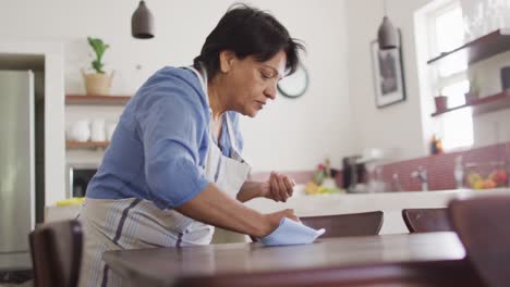 senior biracial woman wearing apron and cleaning table in kitchen alone